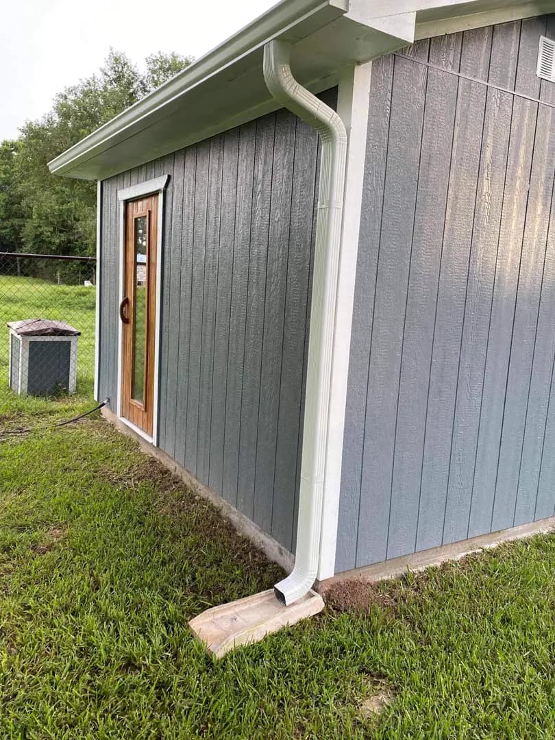 Full white downspout running from the gutter to a concrete splash block on a gray detached building